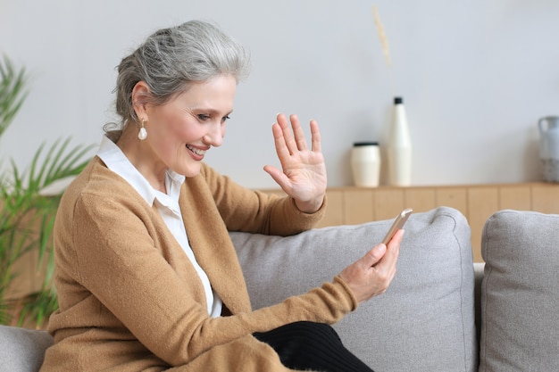 Smiling middle aged woman holding phone, using mobile device apps, looking at screen, while sitting on couch.