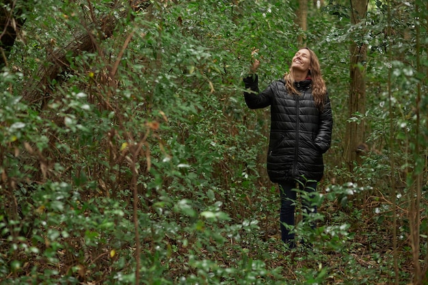Smiling middle-aged woman strolling in the woods in winter, while caressing and enjoying the greenery. Caucasian blonde hair.