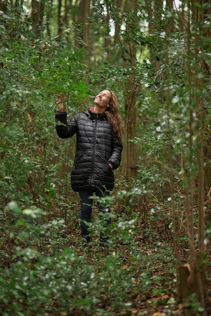 Smiling middle-aged woman strolling in the woods in winter, while caressing and enjoying the greenery. Caucasian blonde hair.