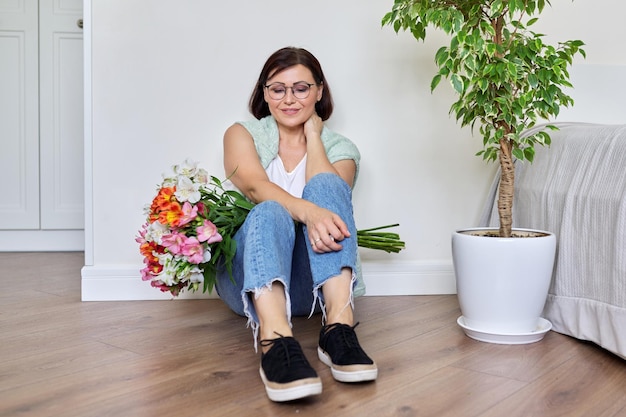 Smiling middle aged woman with bouquet of flowers sitting on the floor at home