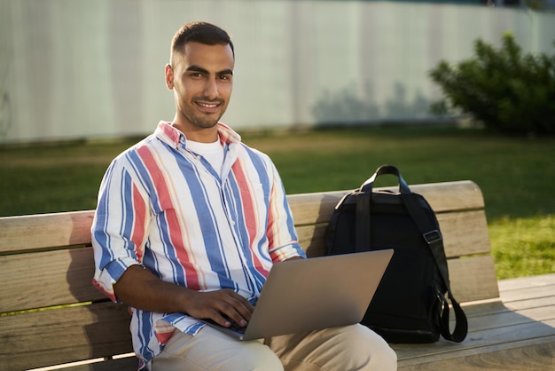 Smiling middle eastern man freelancer using laptop computer working online sitting on bench