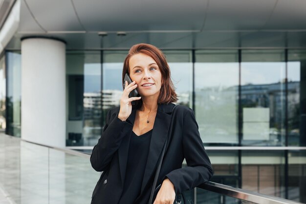 Smiling middleaged business woman with ginger hair leaning on a railing talking on a mobile phone