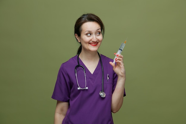 Smiling middleaged female doctor wearing uniform and stethoscope around neck holding syringe with needle looking at side isolated on olive green background