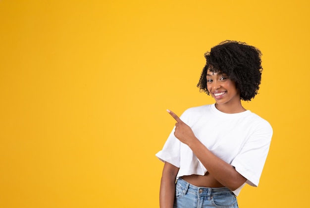 Smiling millennial african american curly woman in white tshirt pointing fingers at empty space