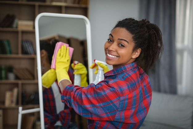 Foto donna afroamericana millenaria sorridente in guanti di gomma che lava lo specchio con spray all'interno della stanza