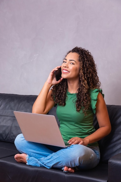 Smiling millennial african american woman talking on the phone using laptop at home office, happy young mixed race lady making call having mobile conversation looking at computer screen sit on sofa