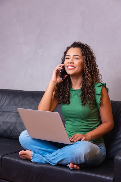 Smiling millennial african american woman talking on the phone using laptop at home office, happy young mixed race lady making call having mobile conversation looking at computer screen sit on sofa