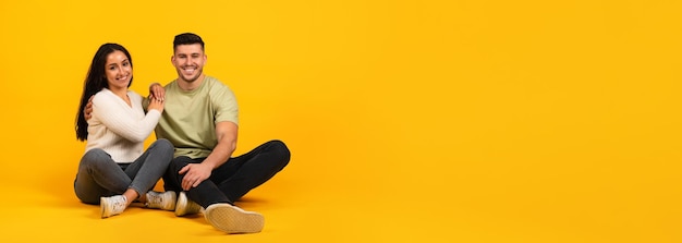 Smiling millennial arabic female and male in casual sit on floor with free space isolated on orange background