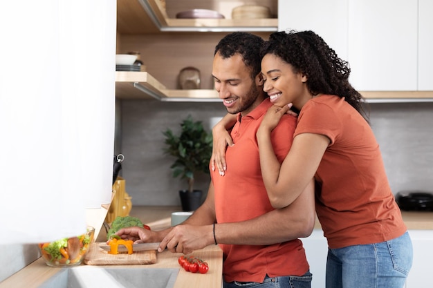 Smiling millennial black wife hugging her husband guy cuts organic vegetables at salad in kitchen interior