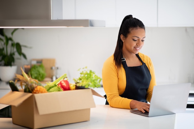 Smiling millennial black woman in apron typing on laptop at table with cardboard box with organic vegetables