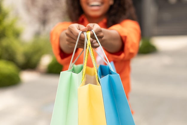 Smiling millennial black woman in casual shows many of bags\
with order enjoys great shopping in city cropped