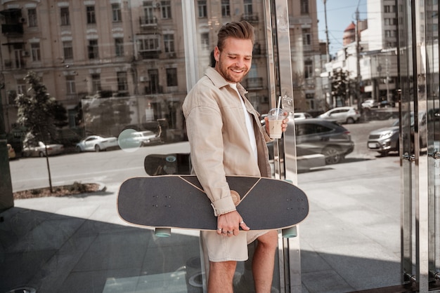 Smiling millennial blonde guy holding cup of coffee cappuccino
takeaway standing on urban street glass building background hipster
skateboarder holding longboard. extreme leisure concept. city
habits.