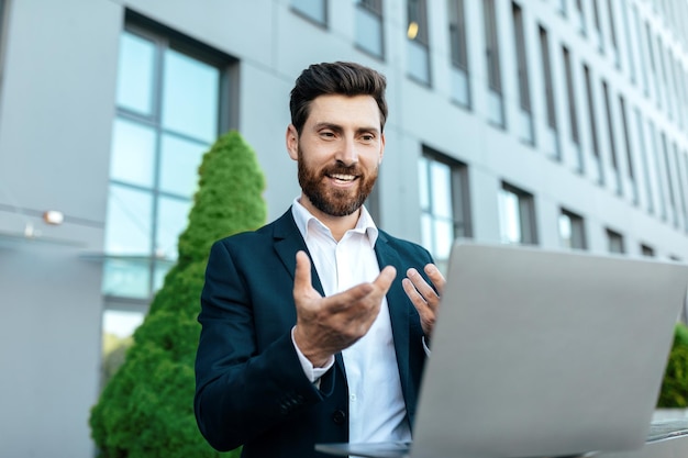 Smiling millennial caucasian businessman in suit with beard working on laptop has video call