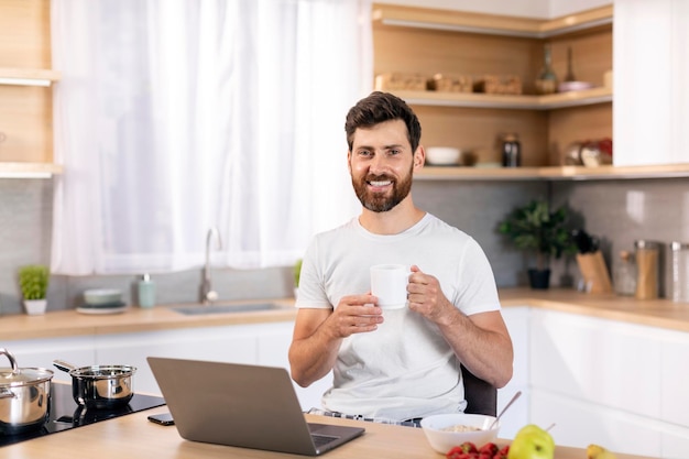 Smiling millennial caucasian male with beard with cup of coffee enjoys good morning has breakfast alone with computer