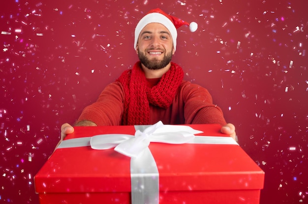 Smiling millennial caucasian man with beard in santa hat give box present with snow confetti