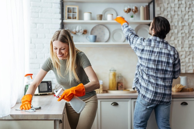 Smiling millennial european woman blonde with rubber gloves and man wipe dust on light kitchen