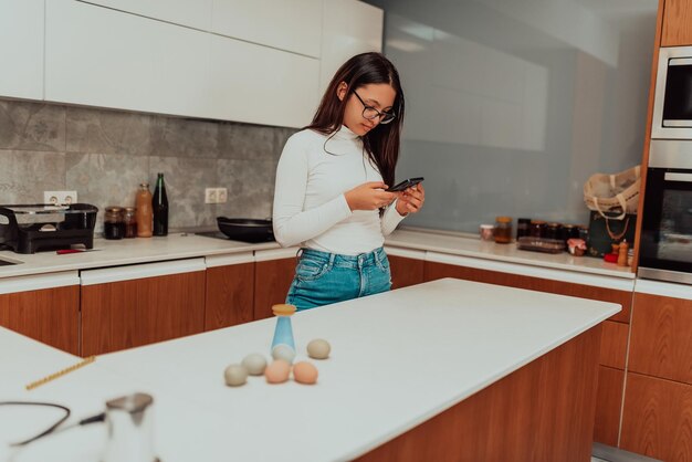 Smiling millennial girl stand at white modern kitchen counter texting messaging on modern cell gadget happy young woman renter using smartphone device with wireless Internet enjoy weekend at home