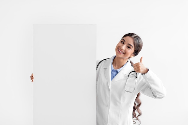 Smiling millennial indian woman doctor in white coat hold big paper banner with blank space and show thumb up