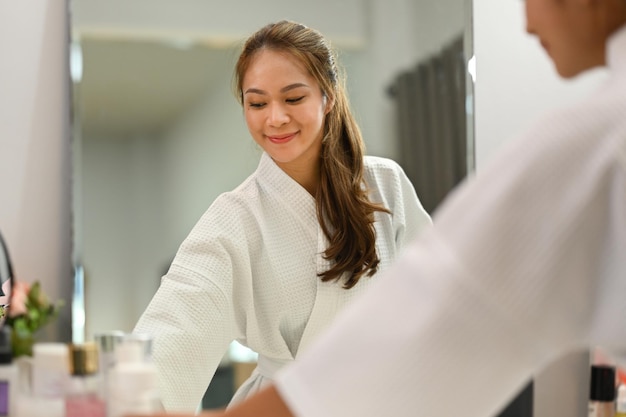 smiling millennial woman in bathrobe standing front of mirror and making daily beauty routine at home