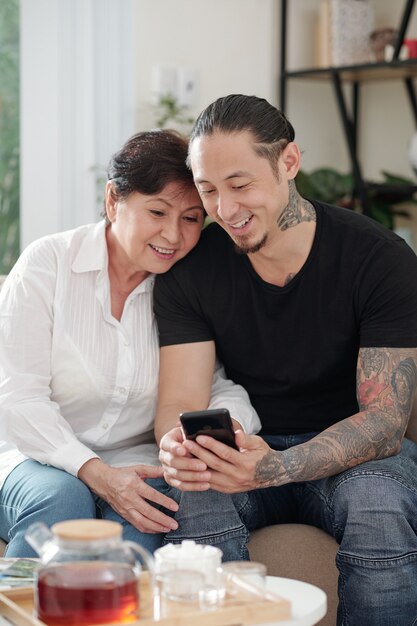 Smiling mixed-race man visiting mother and showing her photos and videos on his smartphone