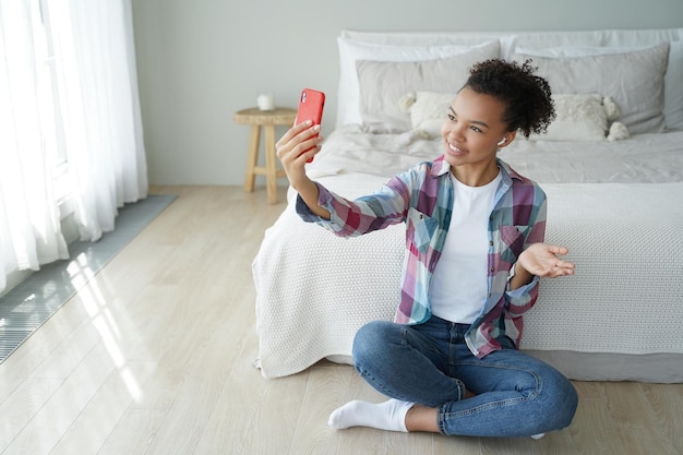 Smiling mixed race young girl blogger talking online by video call sitting on the floor in bedroom