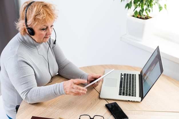 이어폰을 끼고 웃고 있는 현대적인 60대 백인 여성이 집에서 온라인으로 웨비나를 시청합니다. 행복한 수석 회색 머리 여성은 인터넷에서 재미있는 공부를 합니다. 노인 기술 개념입니다.