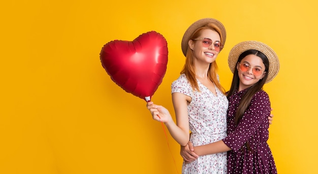 Smiling mom and daughter hold love heart balloon on yellow background