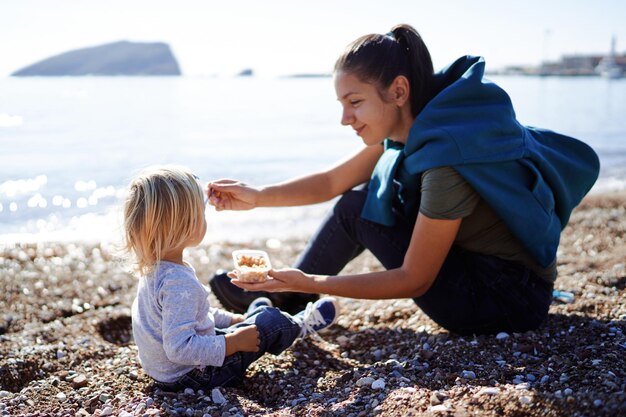 写真 笑顔の母が海辺に座っている間小さな女の子にスプーンでポリッジをあげています