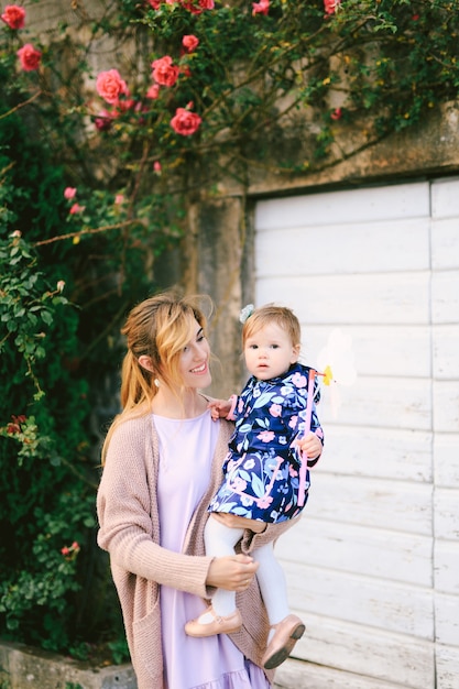 Smiling mom holding little daughter with a toy flower in her arms against the background of a house