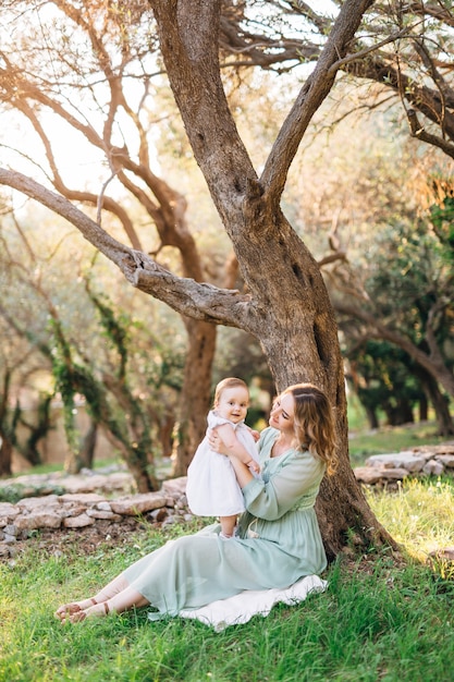 Smiling mom holding a little girl in her arms while sitting under a tree in the park