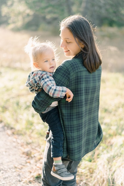 Smiling mom stands in a clearing with a little girl in her arms and looks at her side view