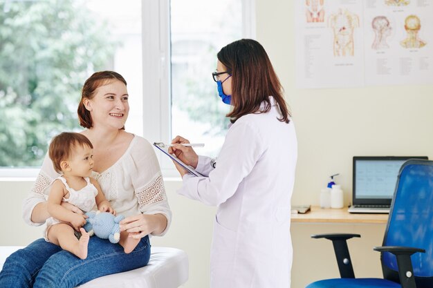 Foto madre sorridente della bambina adorabile che risponde alle domande del pediatra nella mascherina medica