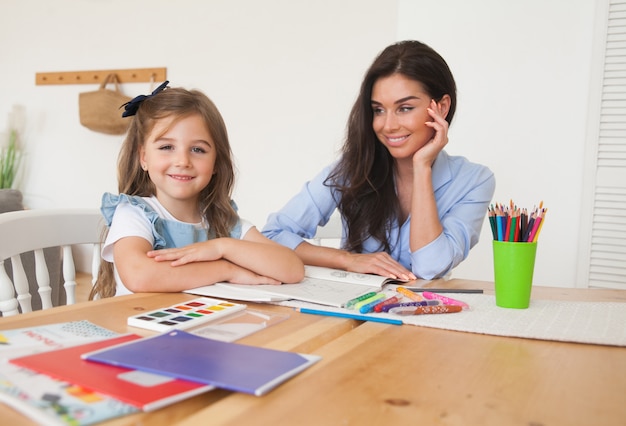 Smiling mother and daughter preparing for lessons and draws at the table with pencils and paints