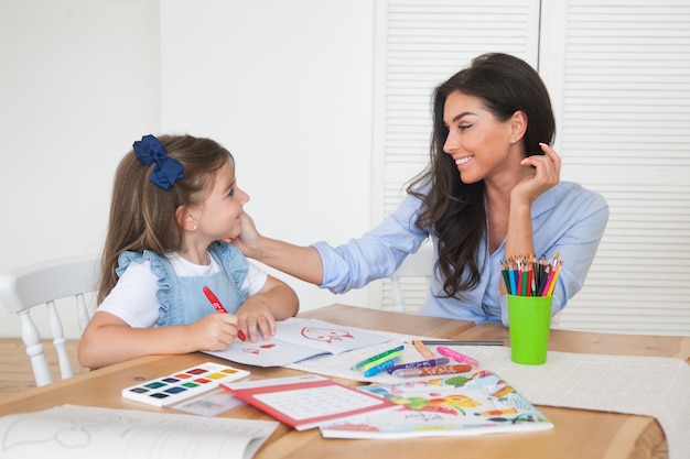 Smiling mother and daughter preparing for lessons and draws at the table with pencils and paints