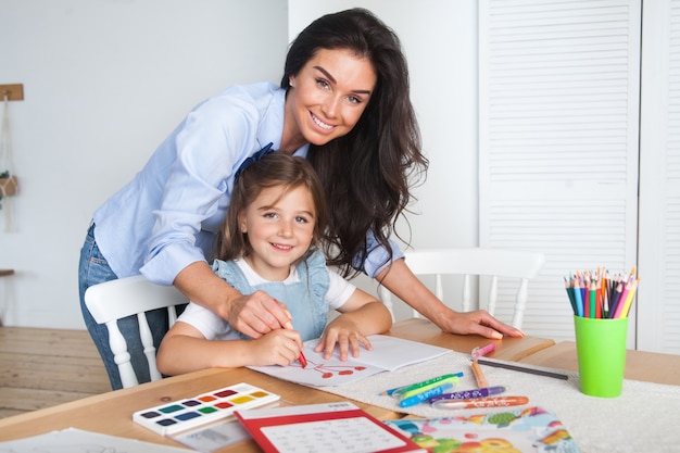 Smiling mother and daughter preparing for lessons and draws at the table with pencils and paints