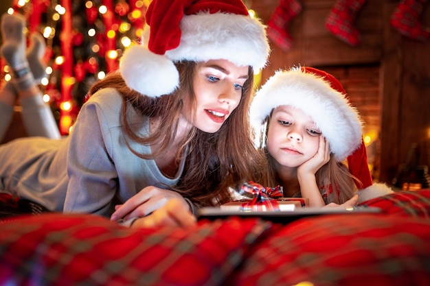 Улыбающаяся мать и дочь в новогодних шапках и пижамах смотрят смешные видеоролики