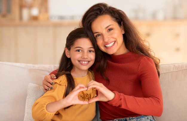 Улыбающиеся мать и дочь показывают символ сердца, сидя дома