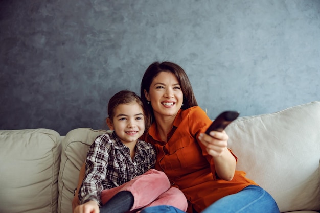 Smiling mother and daughter sitting on the sofa and watching cartoons Mother holding remote control