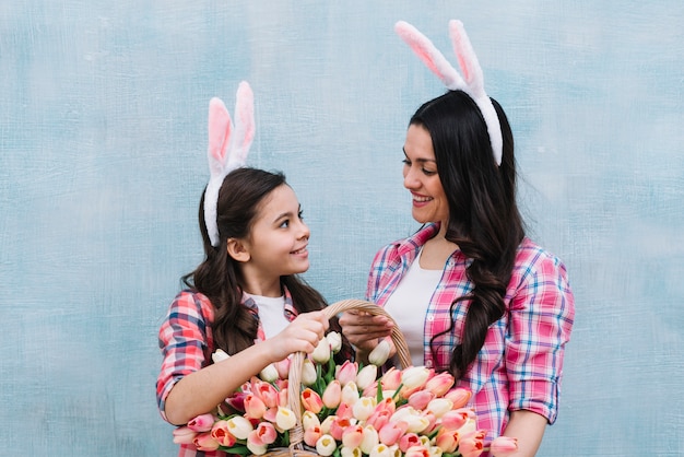 Smiling mother and daughter wearing bunny ears holding tulips basket looking at each other against blue wall