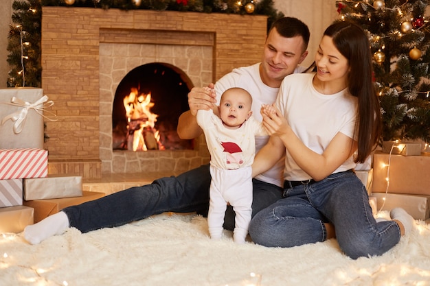 Smiling mother and father spending time together with their infant daughter while sitting on floor near fireplace and X-mas tree, merry Christmas and happy new year.