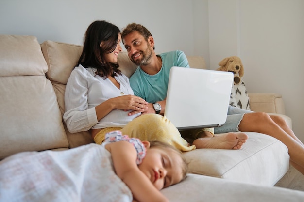 Smiling mother and father watching video on laptop near\
sleeping daughter