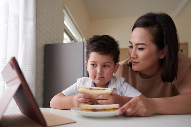 タブレットコンピュータで教育ビデオを見ている幼い息子にサンドイッチを与える笑顔の母親