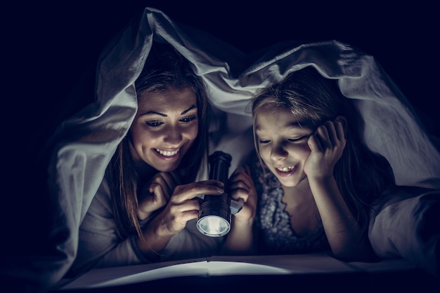 Smiling mother and her daughter reading book under bed cover and holding a flashlight.