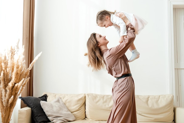 Photo smiling mother lifts happy little daughter into the air in the living room and plays with her