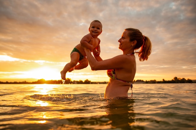 Smiling mother playing holding baby above sea water during sunset.