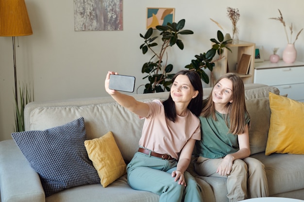 Sorridente madre seduta sul divano nel soggiorno e prendendo selfie con la figlia sullo smartphone