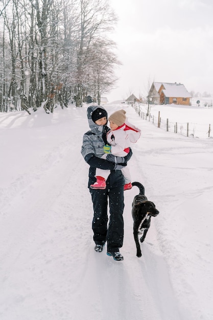 小さな女の子を腕に抱いた笑顔の母親が犬と一緒に雪の降る村の道を歩く