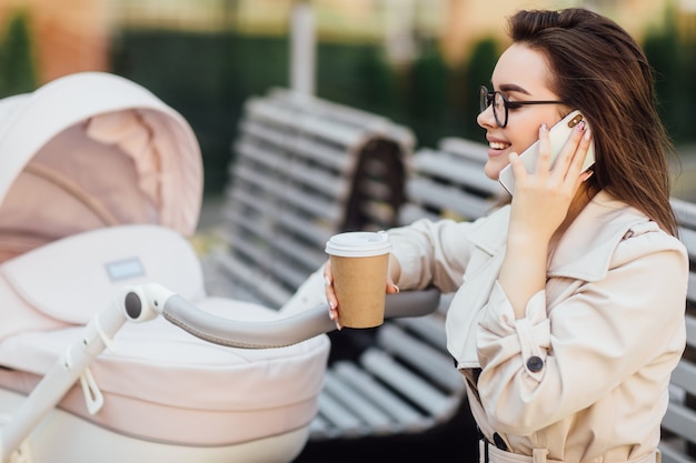 Smiling mother with a newborn baby in a stroller talking by her phone