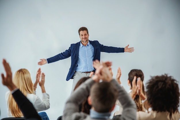Photo smiling motivational speaker standing in front of his audience who is clapping.