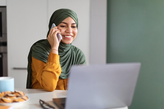 Smiling Mulsim Woman Using Laptop And Talking On Cellphone At Home